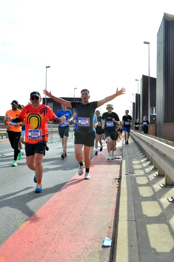 A group of runners participating in a race, with one male runner celebrating with arms outstretched. The scene shows a mix of running gear and a city backdrop.