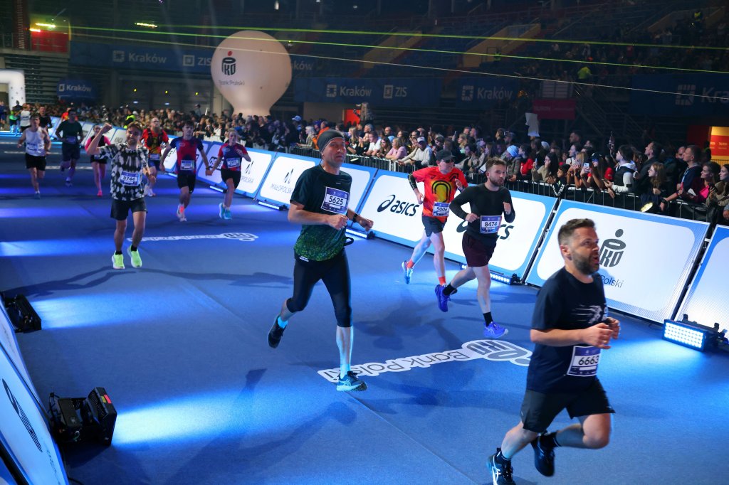 A group of runners participating in a race event indoors, with a cheering crowd in the background. The athletes are wearing numbered bibs and various running gear, highlighted by bright stage lighting.
