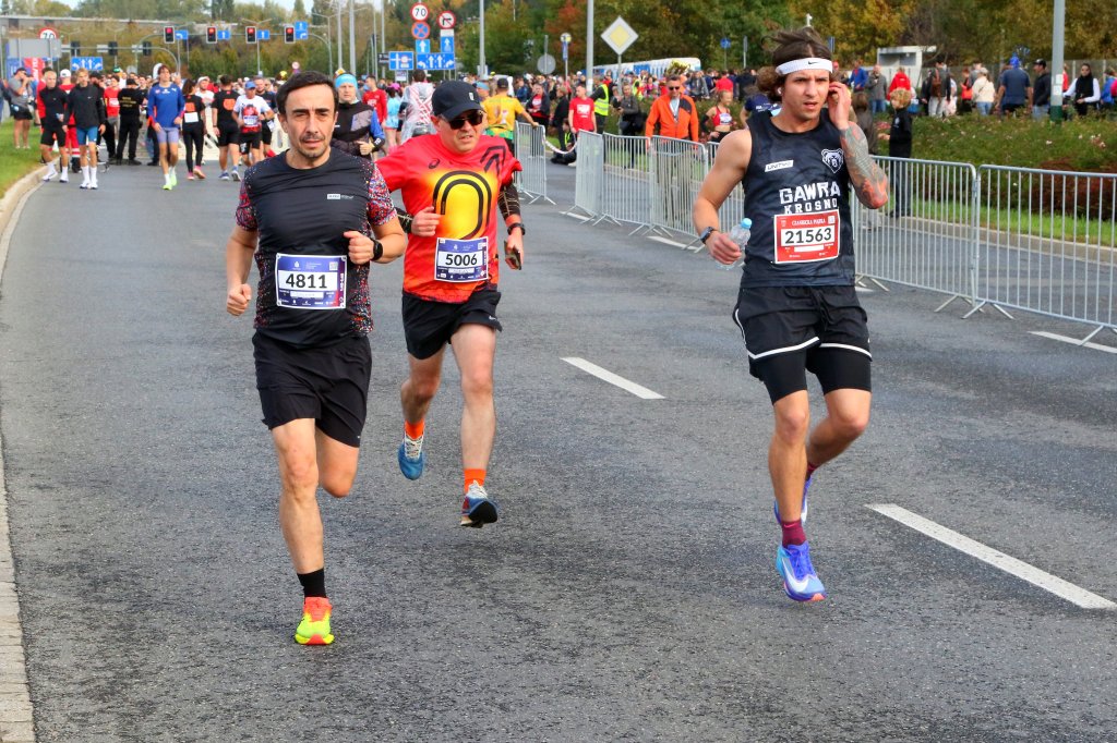 Three runners competing in a marathon on a city street, surrounded by a crowd of participants in the background. The runners wear numbers pinned to their chests and are dressed in athletic gear.