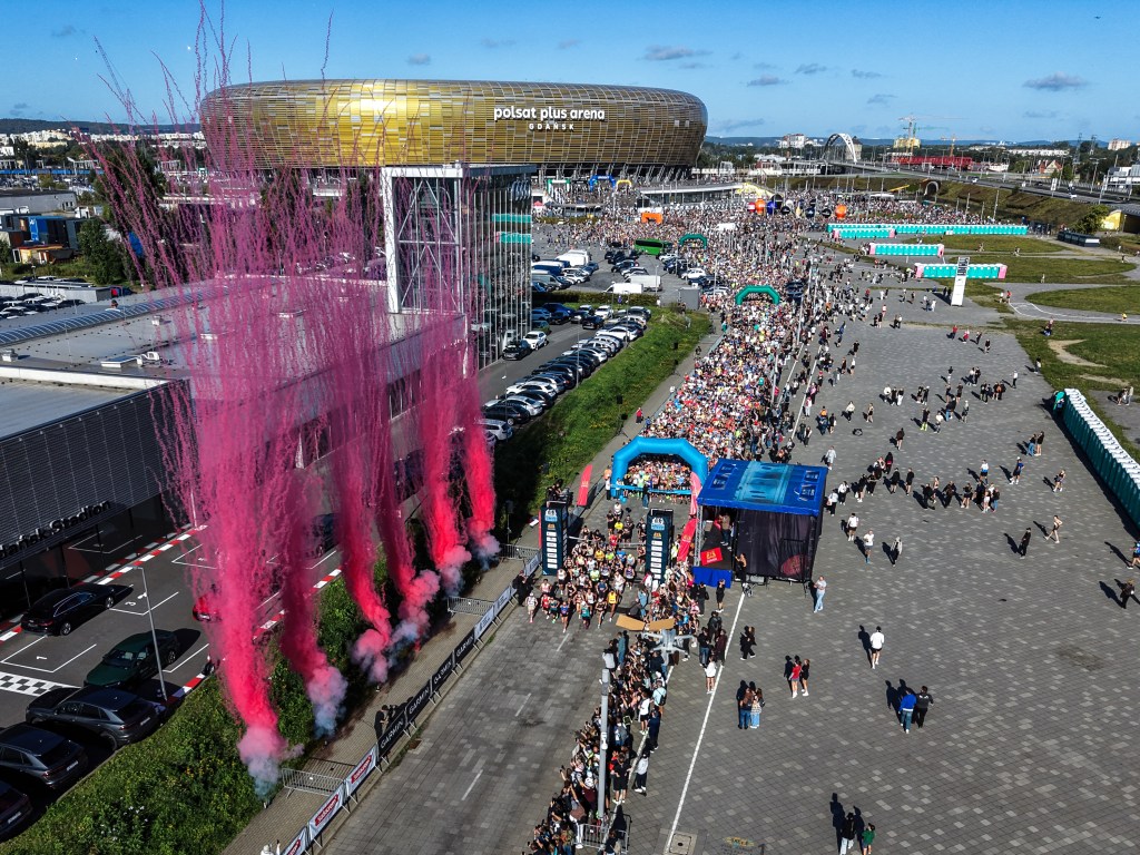Aerial view of Polsat Plus Arena with a large crowd and pink smoke emerging from the building. Multiple people are gathered in line outside the venue, with portable restrooms visible nearby.