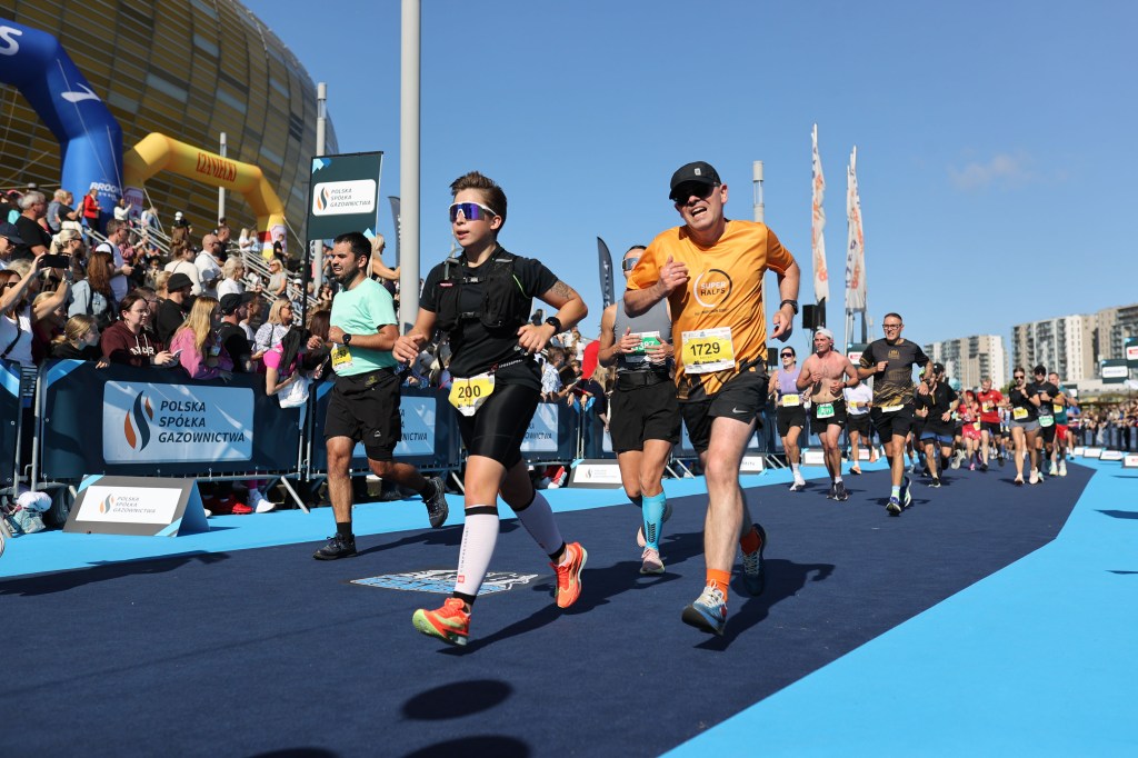Participants running in a race event with a crowd of spectators in the background on a sunny day.